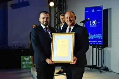 Dwaj mężczyźni w garniturach stoją na scenie i trzymają wspólnie oprawiony w złotą ramę dyplom podczas uroczystości jubileuszu 120-lecia Piekarni Krakowskiego.